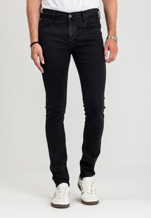 Mężczyzna noszący czarne jeansy typu slim fit w połączeniu z białymi trampkami, białą koszulką, zegarkiem na lewym nadgarstku i gumką do włosów na prawym nadgarstku.