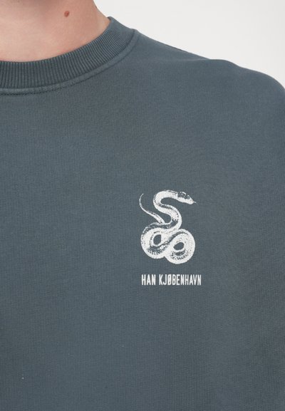 Sötétzöld pulóver, fehér kígyó grafikával és "HAN KJØBENHAVN" felirattal a minta alatt. Puha tapintású, bordázott kerek nyakkal.