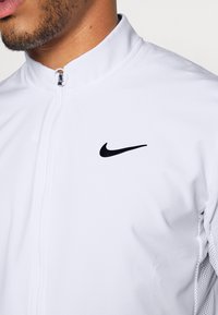 Chaqueta blanca de Nike con un alto cuello, cremallera completa, logo negro en el pecho y detalles texturizados a lo largo de los lados.