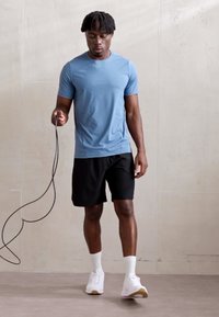 Ανοιχτό μπλε αθλητικό T-shirt, μαύρες βερμούδες και λευκά αθλητικά παπούτσια; κρατάει ένα σχοινάκι. Λείο ύφασμα, εφαρμοστή σχεδίαση, casual στυλ.