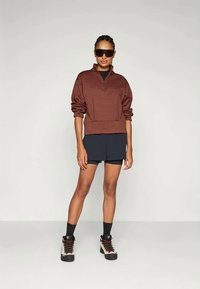 Brun zip-up genser med lange ermer, kombinert med mørke shorts og høye tursko. Modell iført solbriller, står mot en enkel bakgrunn.