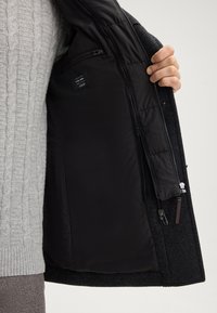 Una mano tiene aperto un cappotto scuro che mostra la fodera interna nera liscia con tasca con zip e etichetta di vestibilità all'interno.