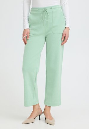 Femme portant un pantalon vert clair à cordon de serrage avec poches avant et des talons slingback beige, debout devant un fond blanc uni.