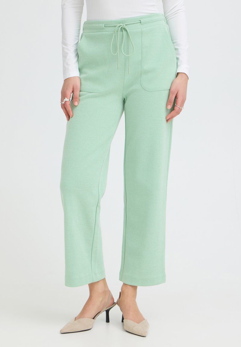 Femme portant un pantalon vert clair à cordon de serrage avec poches avant et des talons slingback beige, debout devant un fond blanc uni.
