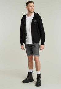 Zwarte zip-up hoodie met voorkeurszakken, gecombineerd met grijze denim shorts en zwarte chunky laarzen. Wit T-shirt eronder, zichtbare branding op de hoodie.