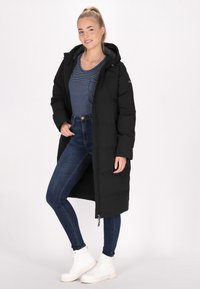 Cappotto lungo nero imbottito con cappuccio, zip frontale e tasche laterali. Sotto, una maglietta a righe blu, abbinata a jeans scuri aderenti e sneakers bianche.