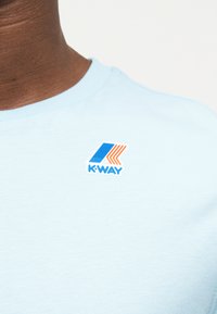 Κοντινό πλάνο ενός ατόμου που φοράει ανοιχτό μπλε πουκάμισο με λογότυπο K-Way σε μπλε και πορτοκαλί στο στήθος.