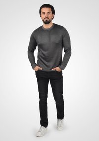 Suéter gris texturizado con cuello redondo, combinado con jeans negros y zapatillas blancas. El modelo está de pie con las manos en los bolsillos sobre un fondo claro.