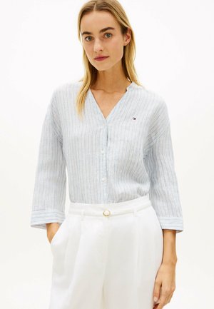 Frau mit blondem Haar, die ein hellblau gestreiftes Hemd mit Knöpfen und hoch taillierte weiße Hose mit dünnem Gürtel vor weißem Hintergrund trägt.