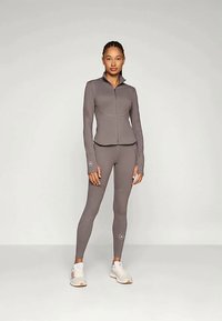 Grå atletisk outfit bestående af en tætsiddende zip-up jakke og matchende leggings. Glat tekstur med udskæringer på ærmerne.