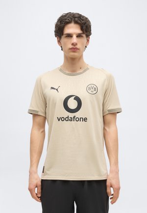 Ung mann iført beige Puma Borussia Dortmund fotballdrakt med Vodafone-logo, stående foran hvit bakgrunn.