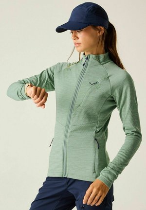 Vrouw in een groene jas met rits en een marineblauwe pet controleert een smartwatch om haar linkerpols, draagt marineblauwe broek.