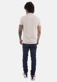 Hombre con tatuajes que lleva una camiseta tipo polo beige de manga corta, jeans azul oscuro y zapatillas blancas, de pie y de espaldas sobre un fondo blanco.