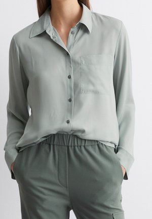 Femme portant une chemise gris clair boutonnée avec une poche poitrine et un pantalon gris foncé à taille élastique, les mains dans les poches.
