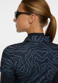 Koszulka sportowa z czarnego materiału, z geometrycznym niebieskim wzorem, krótkimi rękawami i w designie z kołnierzykiem. Na nosie okulary przeciwsłoneczne.