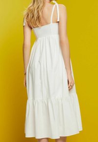 Femme blonde portant une robe blanche sans manches avec des bretelles nouées aux épaules, debout devant un fond jaune moutarde uni.