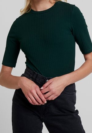 Vrouw die een donkergroen geribbeld kort mouwtopje draagt en een zwarte high-waist spijkerbroek, met haar handen in elkaar gevouwen op haar taille.