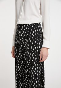 Schwarze gemusterte Hose mit weißen vertikalen Streifen, ergänzt durch eine leichte weiße Bluse mit Knopfdesign.