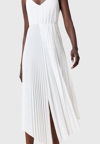 Femme portant une robe midi plissée blanche sans manches avec un col en V et un ourlet asymétrique sur un fond uni.