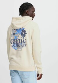 Sweatshirt à capuche couleur crème avec un design floral dans des tons bleus et foncés, avec le texte "GROW AS YOU GO" au dos.