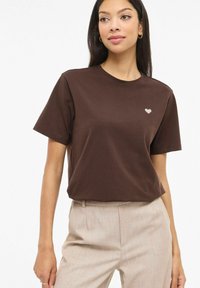 T-shirt di cotone marrone con maniche corte e scollo rotondo, caratterizzata da un piccolo ricamo a forma di cuore chiaro sul petto sinistro.