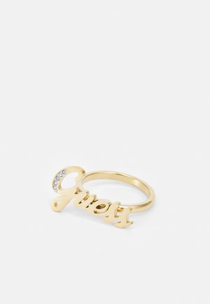 Bague dorée avec le logo "Guess" en écriture manuscrite et petits strass transparents sur la lettre "G", présentée sur fond blanc.