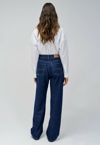 Jeans in denim scuro a gamba larga con vita alta, dotati di tasche posteriori e un'etichetta in pelle marrone del marchio. Abbinati a una camicia bianca con bottoni.