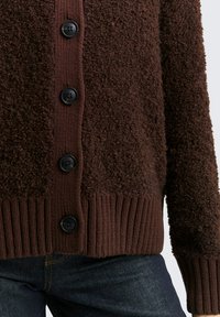 Bruine getextureerde cardigan met een fuzzy oppervlak, met een geribde zoom en zes zwarte knopen aan de voorkant. Draagt over donkere jeans.