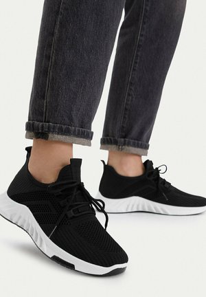 Persona que lleva zapatillas de punto negras con suelas blancas y cordones negros, combinadas con vaqueros oscuros remangados contra un fondo liso.