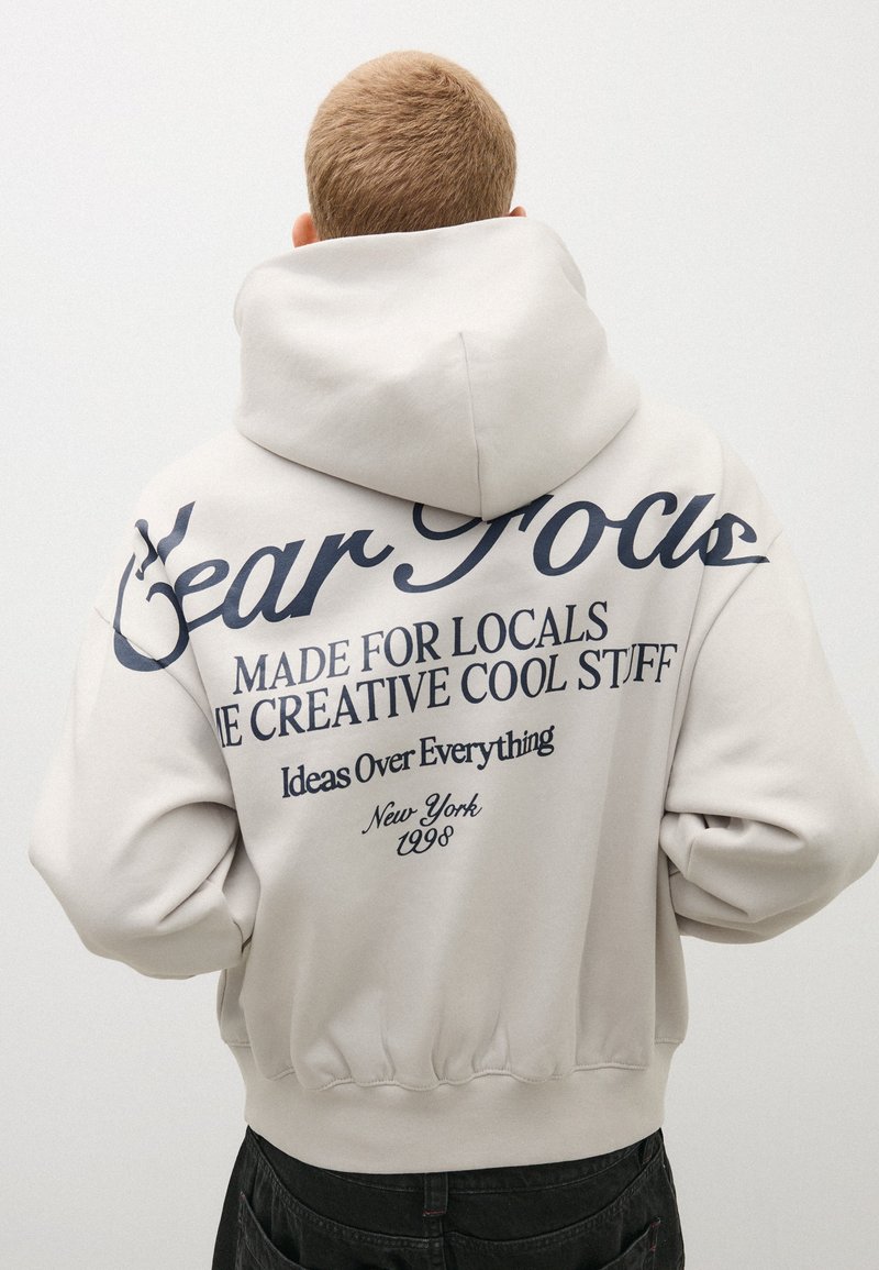 Personne aux cheveux blonds courts portant un sweat à capuche écru avec un texte bleu foncé dans le dos à propos de la créativité et de New York 1998.