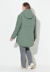 Grön, lättviktsjacka med huva och längre bakdel, sidopanels och en orange logotypaccent. Mjuk textur, avslappnad stil för lager-på-lager.