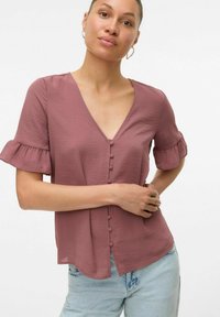 Blusa a maniche corte con scollo a V in rosa polveroso, caratterizzata da un'apertura frontale con bottoni, maniche arricciate e tessuto leggero. Abbinata a jeans azzurri.
