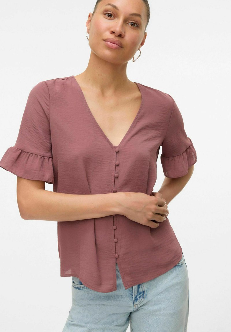 Blusa a maniche corte con scollo a V in rosa polveroso, caratterizzata da un'apertura frontale con bottoni, maniche arricciate e tessuto leggero. Abbinata a jeans azzurri.