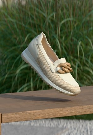 Beige Halbschuhe zum Hineinschlüpfen mit gewebter Textur, Gummisohle und einem verknoteten Akzent vorne. Im Hintergrund grünes Gras.