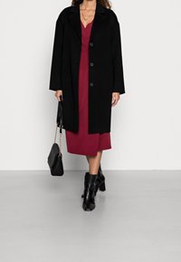 Manteau en mélange de laine noir avec un col à revers et une fermeture à bouton, porté sur une robe bordeaux profonde. Les accessoires incluent un sac à main noir et des bottes à talons.
