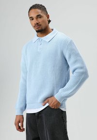 Man met gevlochten haar die een lichtblauwe, gebreide trui met drie knopen, een wit ondershirt en een donkere broek draagt, staand tegen een effen achtergrond.