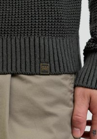 Suéter de punto gris oscuro con dobladillo y puños acanalados, que presenta un patrón texturizado y un pequeño parche de logo en negro y dorado.