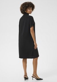 Melna kleita ar taisnu griezumu, ar mežģīņu apdari uz īsām piedurknēm un garumu līdz vidus kājai; valkāta ar melniem siksnām uz papēžiem. Gluda tekstūra.