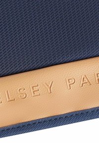 Un tessuto testurizzato blu navy si combina con un dettaglio in ecopelle color beige caratterizzato da un marchio impresso. Il design ha una forma rettangolare con dettagli di cucitura.