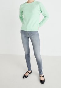 Person i mintgrön tröja, ljusgrå skinny jeans och svarta Mary Jane-skor som står mot en vit bakgrund.