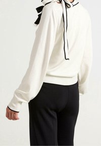 Pull crème avec bordure noire, à manches longues et détail nœud au niveau de la nuque. Associé à un pantalon noir.