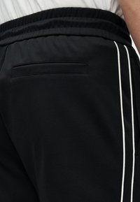 Κοντινό πλάνο μαύρου αθλητικού παντελονιού με λευκές διπλές πλευρικές ρίγες και πίσω τσέπη με φερμουάρ, φορεμένο με λευκό τοπ.