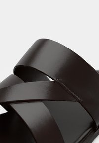 Dark brown leather sandal featuring two crisscrossing straps, a smooth texture, and a flat sole, emphasizing minimalist design.