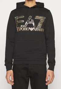 Svart hoodie i mjukt tyg med en glänsande gul och silver logotyp "EA7 Emporio Armani" över bröstet. Framlomma med ficka.