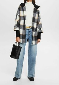 Rutigt överdrag i vitt, grått och svart, med huva och öppen front, kombinerat med en vit polotröja, blåjeans och en svart väska.