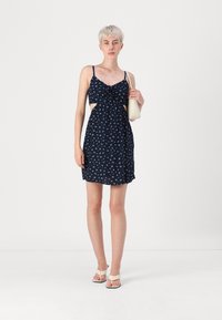 Vestido azul marinho com padrão floral branco, laterais recortadas, frente atada e alças ajustáveis. Usado com uma bolsa bege e sandálias.