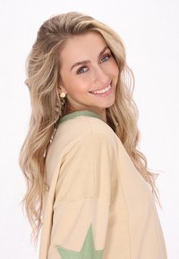 Camicia oversize beige chiaro con un design di stella verde sulla manica sinistra, caratterizzata da un colletto verde a coste e un tessuto morbido e testurizzato.
