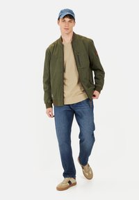 Olivengrøn bomberjakke med ribbede manchetter, beige T-shirt, blå jeans og tan sneakers. Kappen er lyseblå og fuldender det afslappede look.