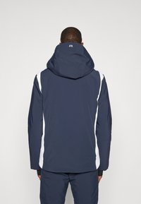 Tengerészkék vízálló dzseki kapucnival, fehér oldalsó kiegészítőkkel és logóval a háton. Sima textúra és sportos szabás.