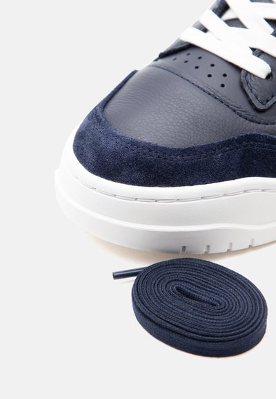 Tmavě modrá teniska s bílou podrážkou a tkaničkami, zobrazující texturovanou kůži a detaily z semiše, s blízkou stočenou tmavě modrou tkaničkou.